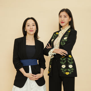 Sara Jane Ho, dressed in semi-formal black blazer with white blouse and Annie Ho dressed in black slacks and a floral patterned blazer, stand confidently against a plain, nude background.